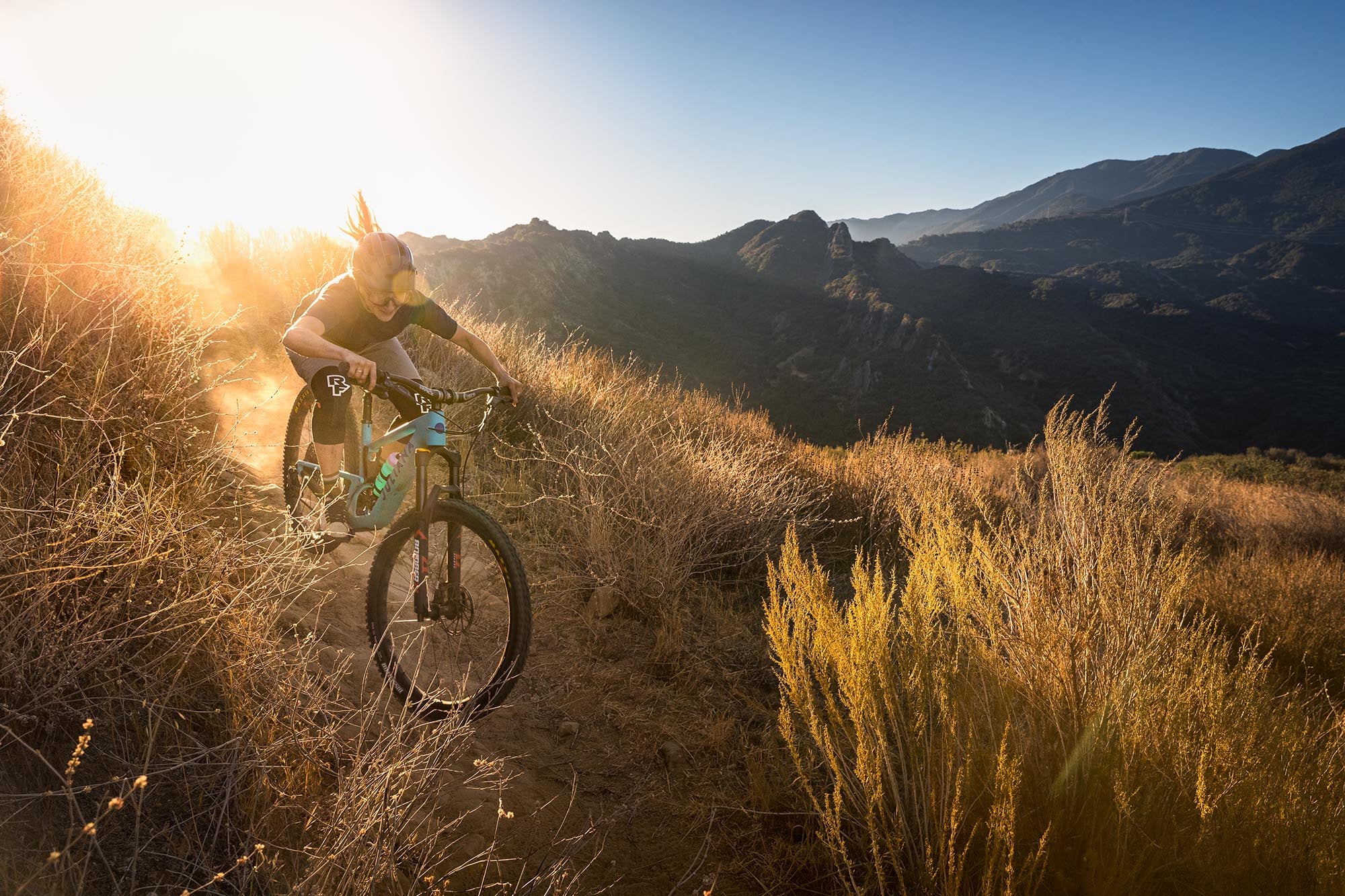 An action shot of a woman riding a Juliana Joplin mountain bike through tall grass, with majestic mountains rising in the background, showcasing the bike’s versatility and performance in natural terrain, available at Contender Bicycles.