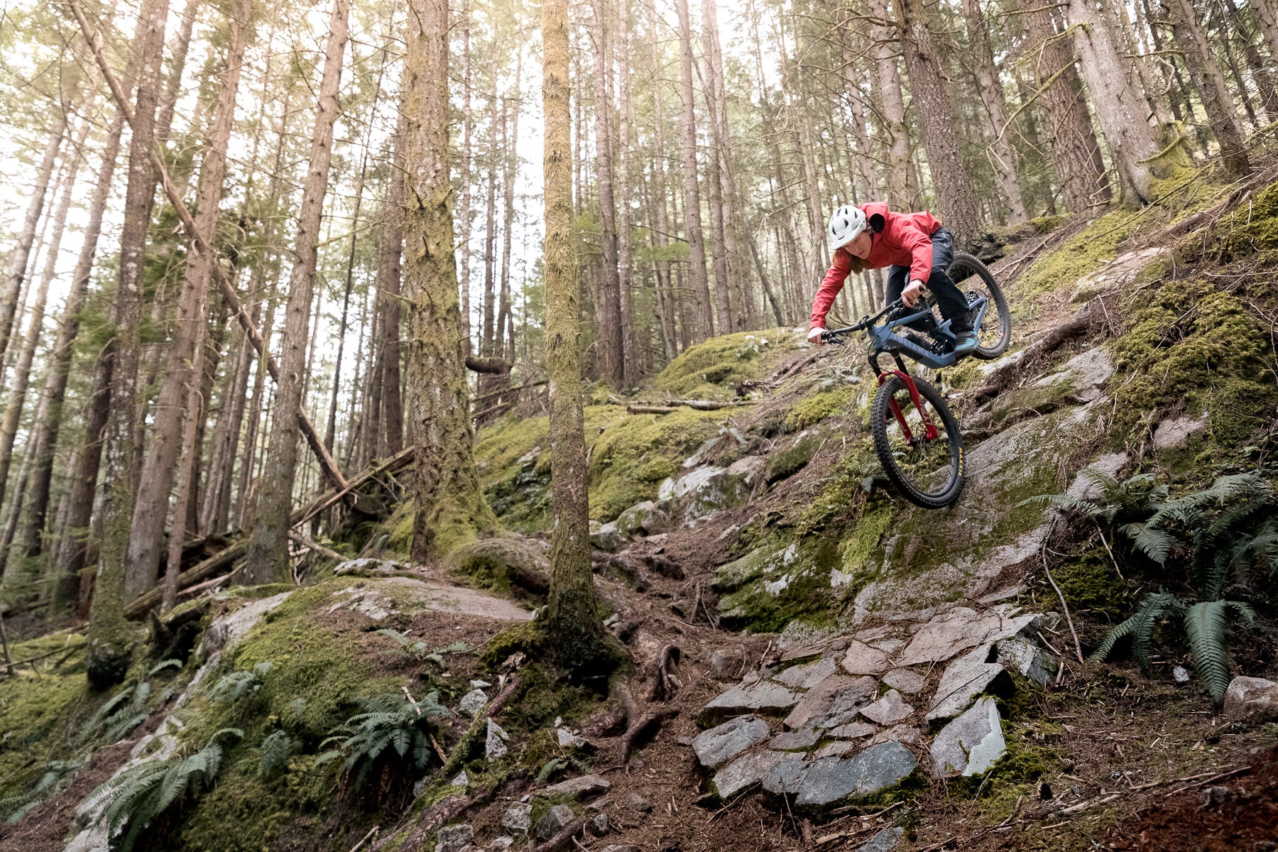 An action shot of a woman riding a Juliana Roubion mountain bike through a dense forest trail, maneuvering around trees and rough terrain, highlighting the bike’s stability and performance in challenging conditions, available at Contender Bicycles.