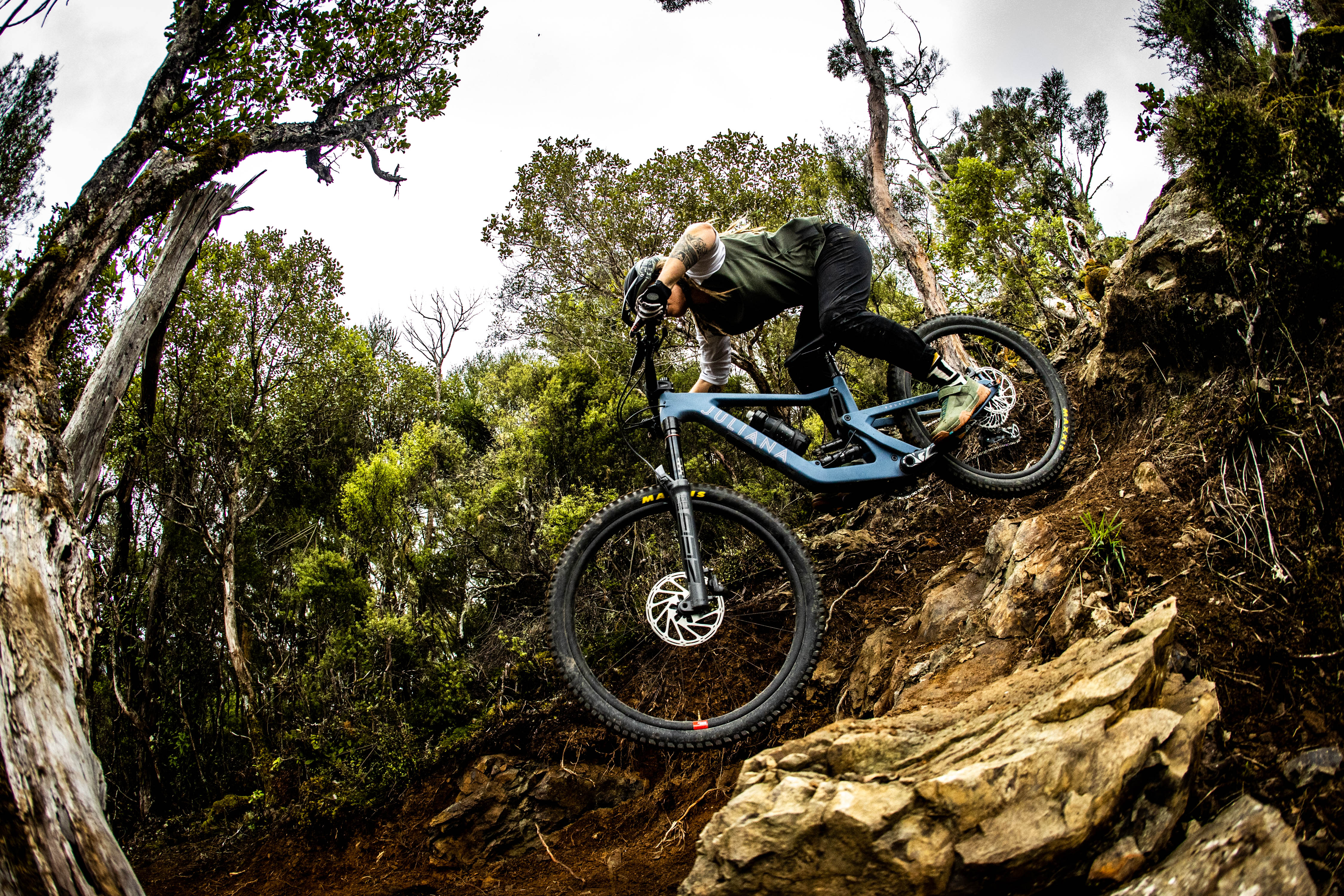 An action shot of a woman riding a Juliana Roubion mountain bike on a scenic trail, surrounded by tall grass and framed by majestic mountains in the background, highlighting the bike’s versatility and performance in rugged terrain, available at Contender Bicycles.
