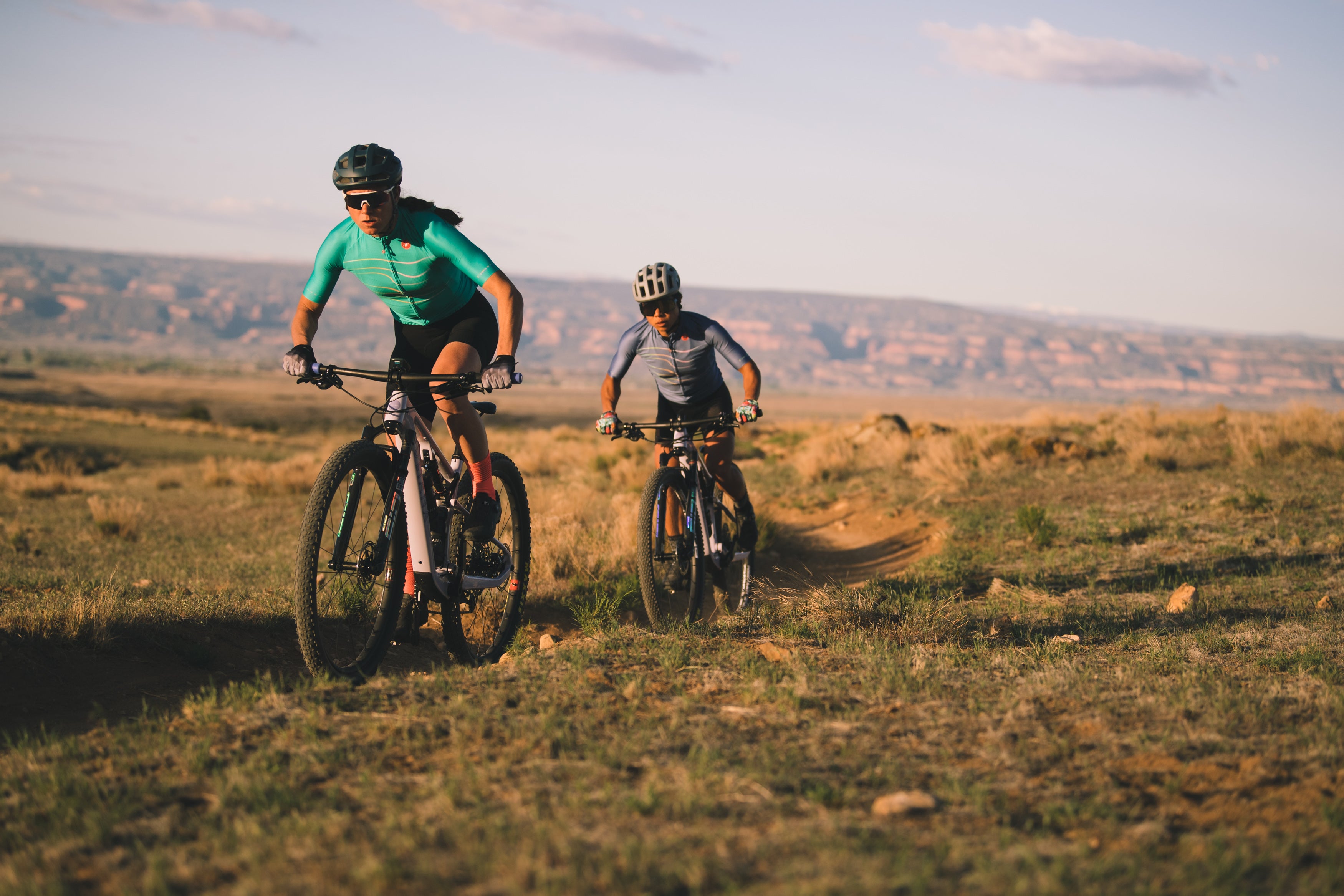 An action shot of two women riding Juliana mountain bikes together on a scenic trail, enjoying the ride and showcasing the bikes' smooth performance on diverse terrain, available at Contender Bicycles.