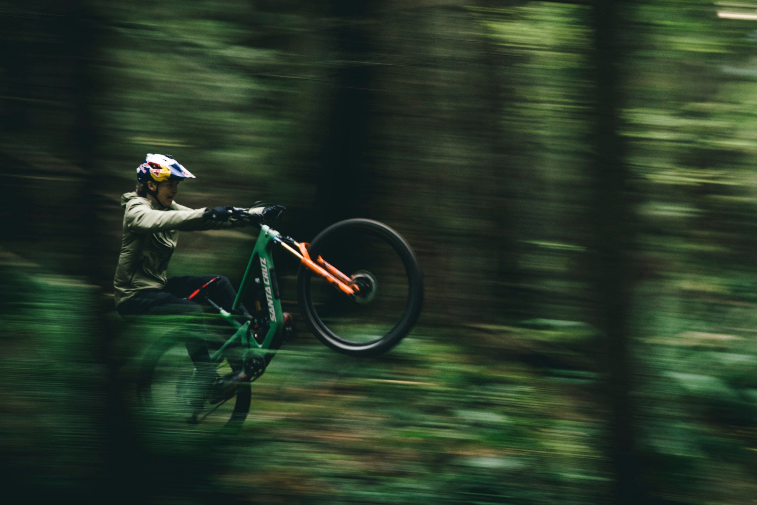 An action shot of a man performing a wheelie on a Santa Cruz 5010 mountain bike, surrounded by towering trees on a scenic forest trail, available at Contender Bicycles.