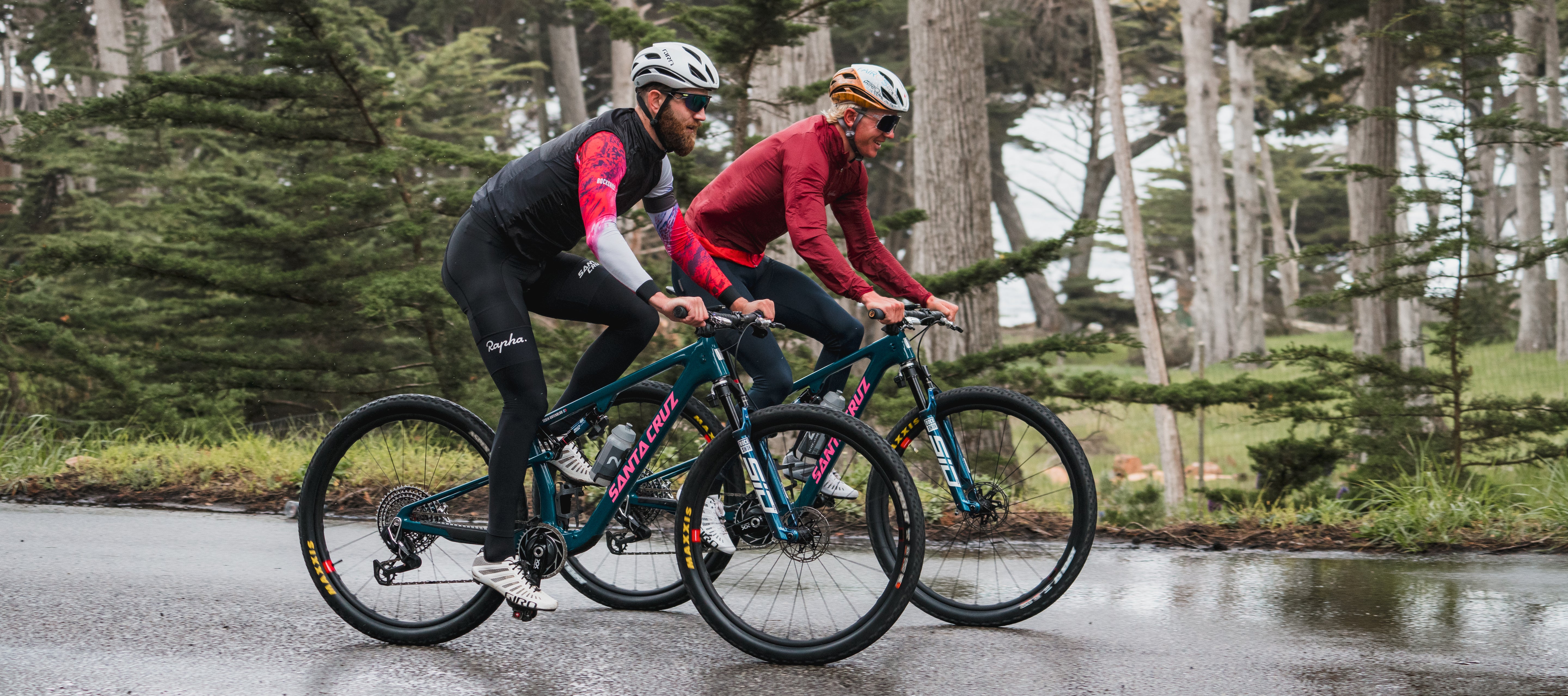 An action shot of two men riding together on Santa Cruz Blur mountain bikes through a lush forest trail, surrounded by towering trees and dappled sunlight, available at Contender Bicycles.