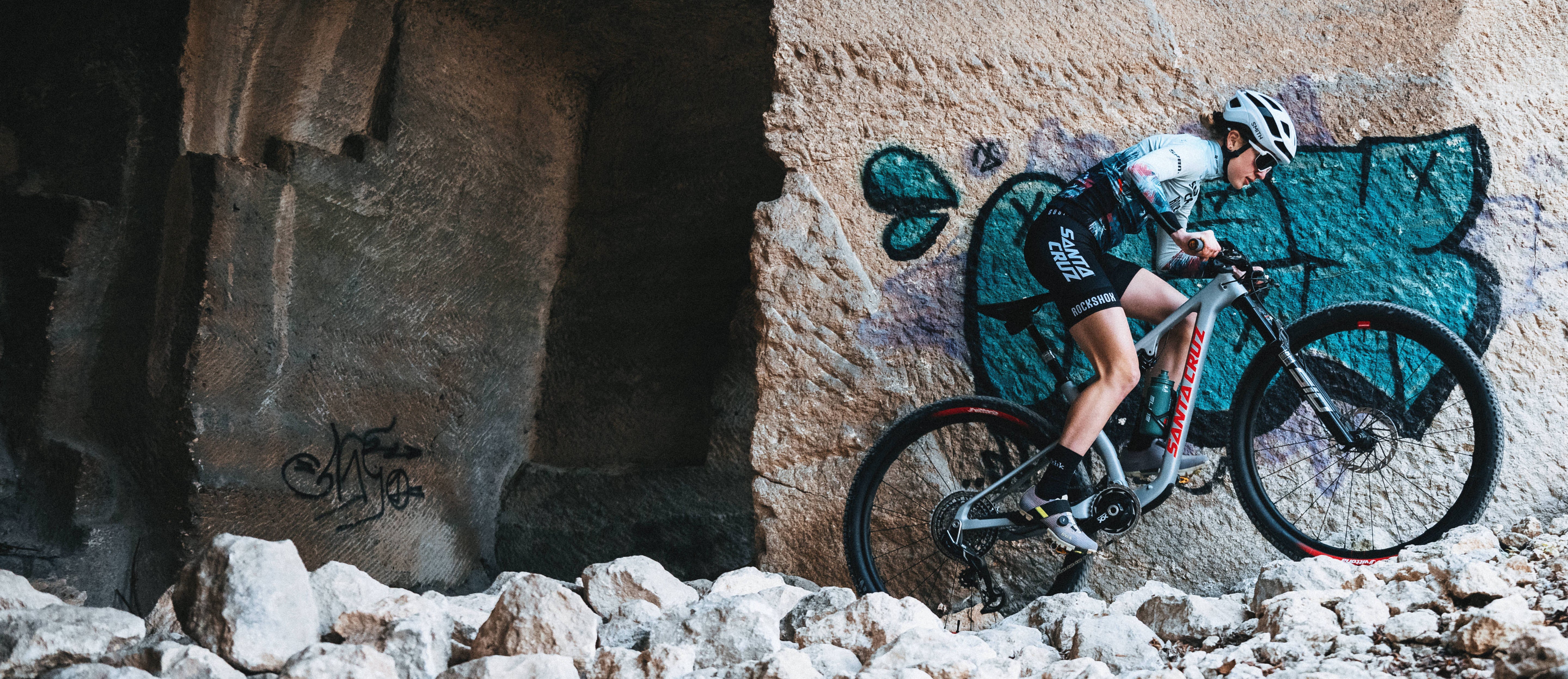 An action shot of a woman riding a Santa Cruz Blur mountain bike over rocky terrain, with vibrant graffiti in the background, showcasing her skill and the bike's performance, available at Contender Bicycles.