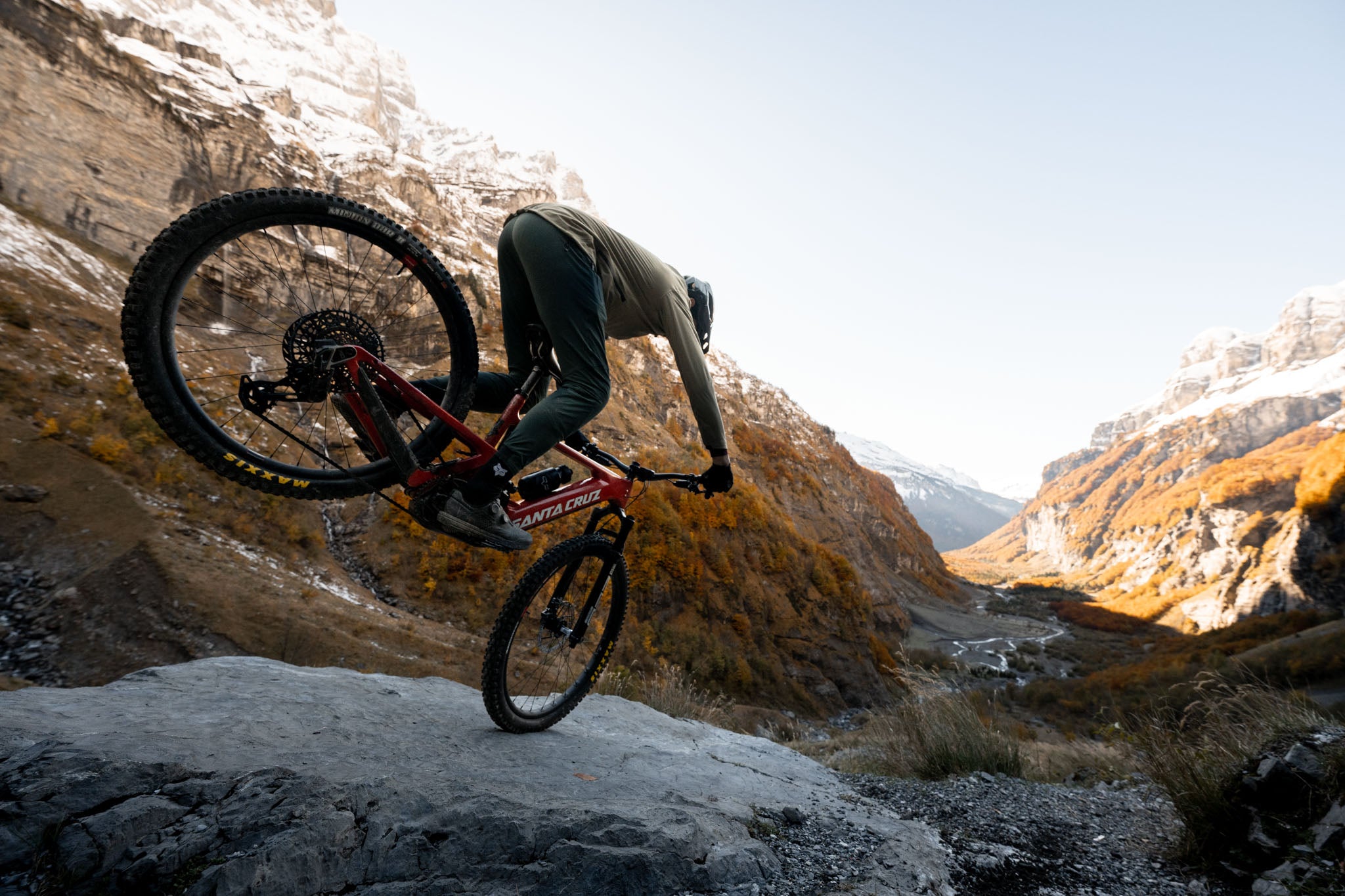 An action shot of a man descending a rainy, rocky mountain trail on a Santa Cruz Heckler SL e-bike, with water splashing and mist swirling around, highlighting the bike's durability and performance in challenging conditions, available at Contender Bicycles.