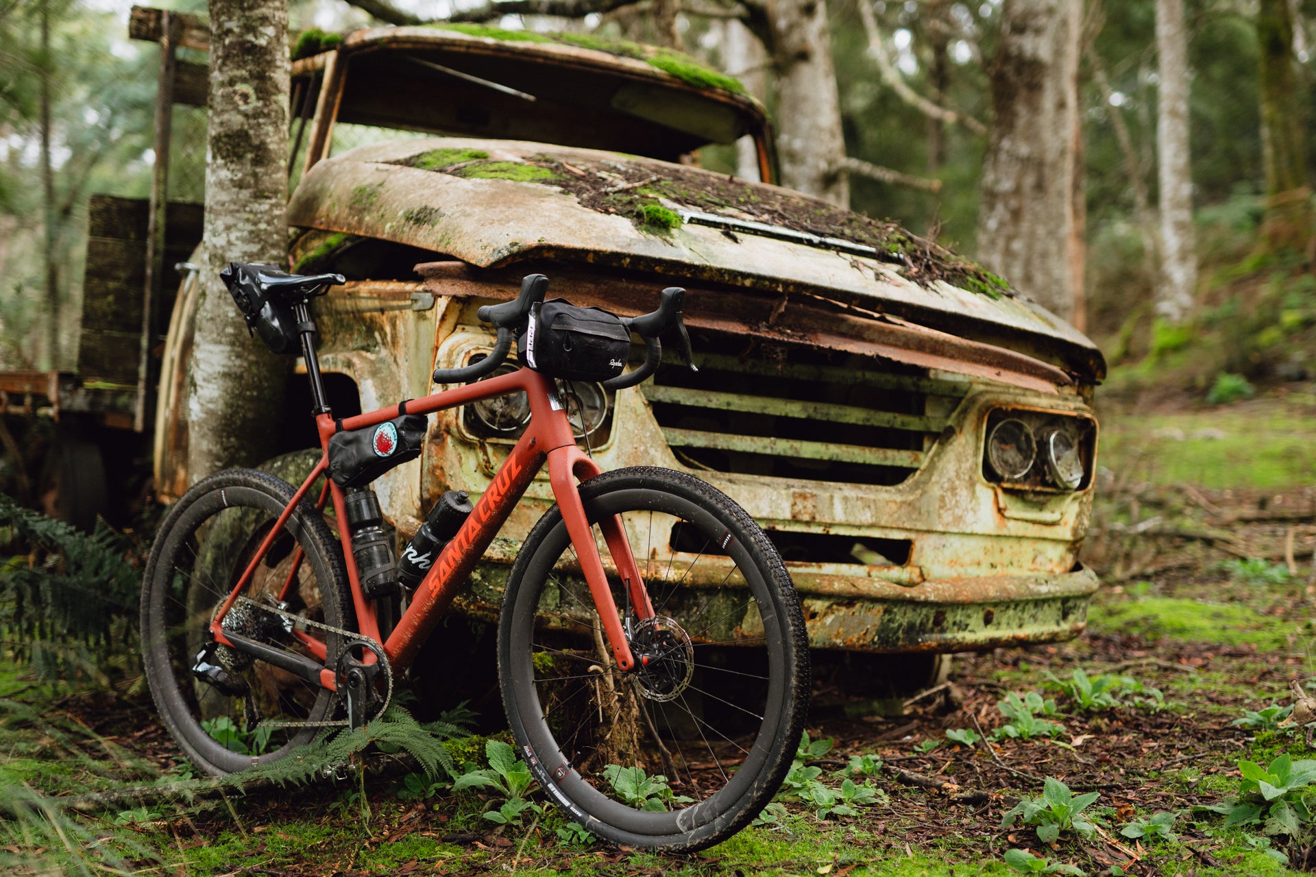 A Red Santa Cruz Stigmata Gravel bike sold at Contender Bicycles, leaned against a rusty truck, photographed in a forest. 