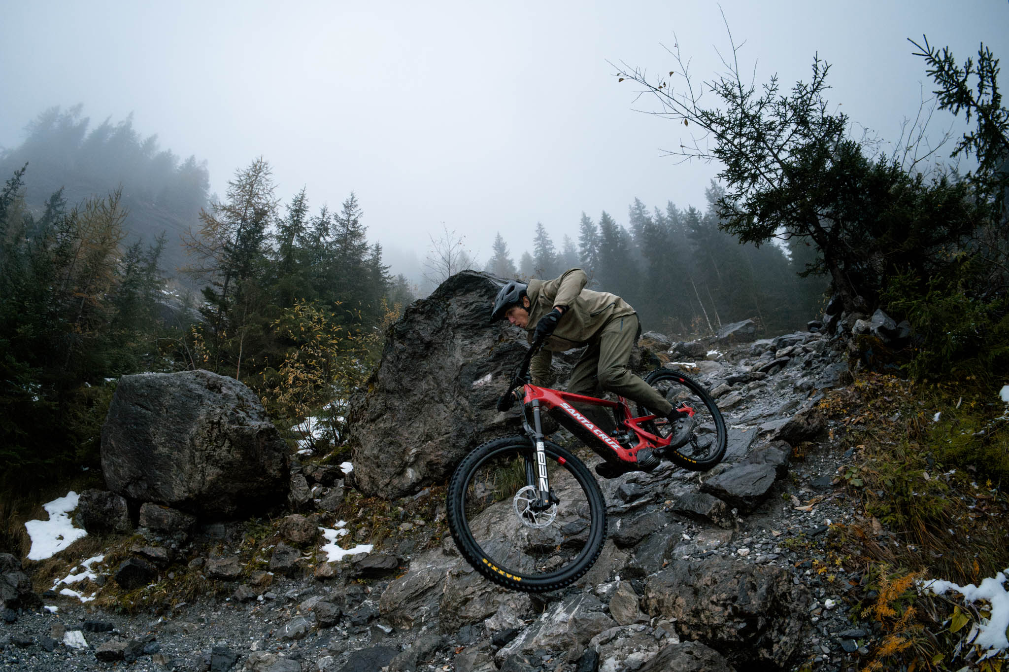 An action shot of a man riding a Santa Cruz Heckler SL e-bike down a rainy, rocky mountain trail, with water splashing and mist in the air, showcasing the bike's stability and performance in tough conditions, available at Contender Bicycles.