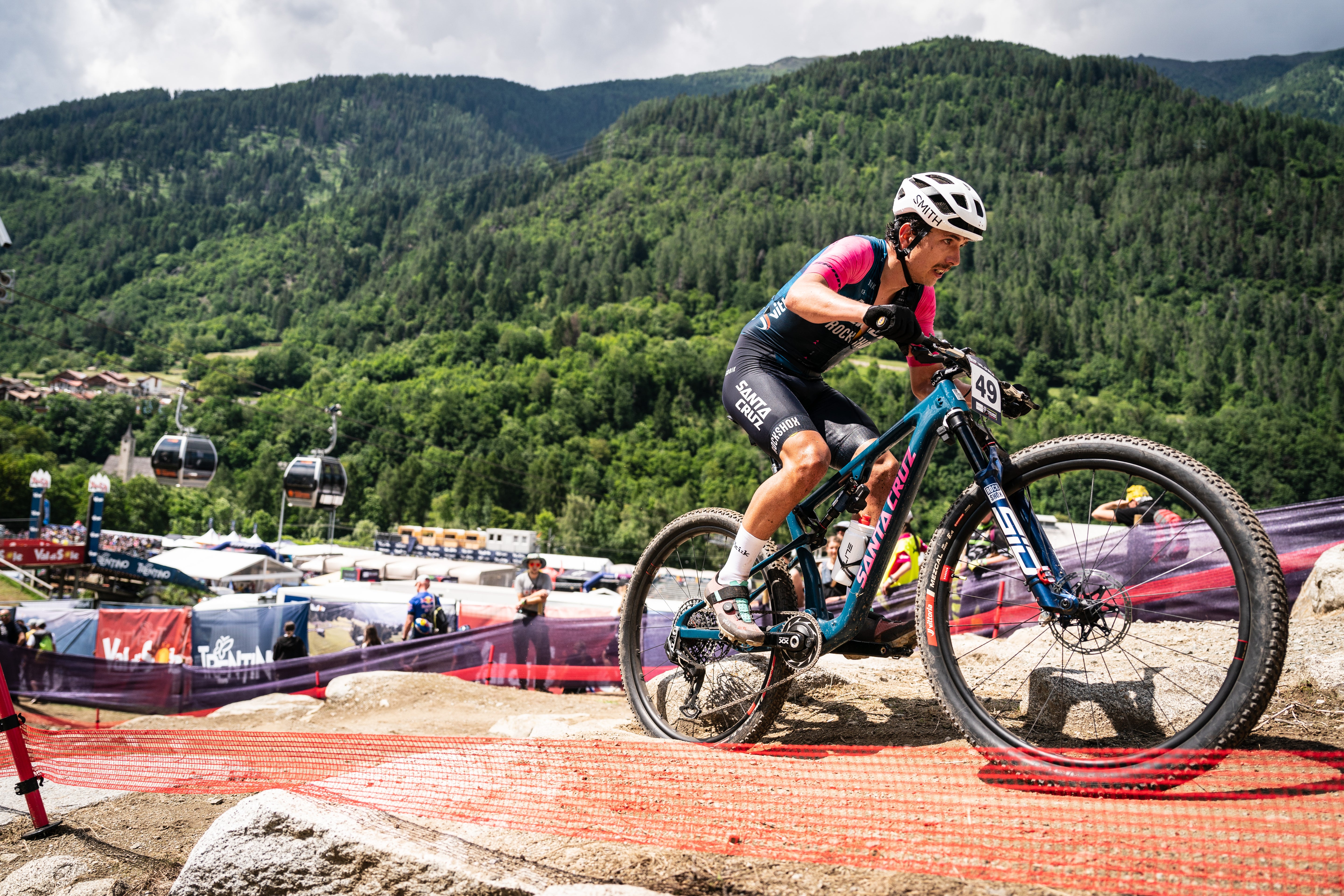 An action shot of a man racing uphill on a Santa Cruz Blur mountain bike, tackling a steep, rocky mountain trail , set against a backdrop of rugged peaks, available at Contender Bicycles.