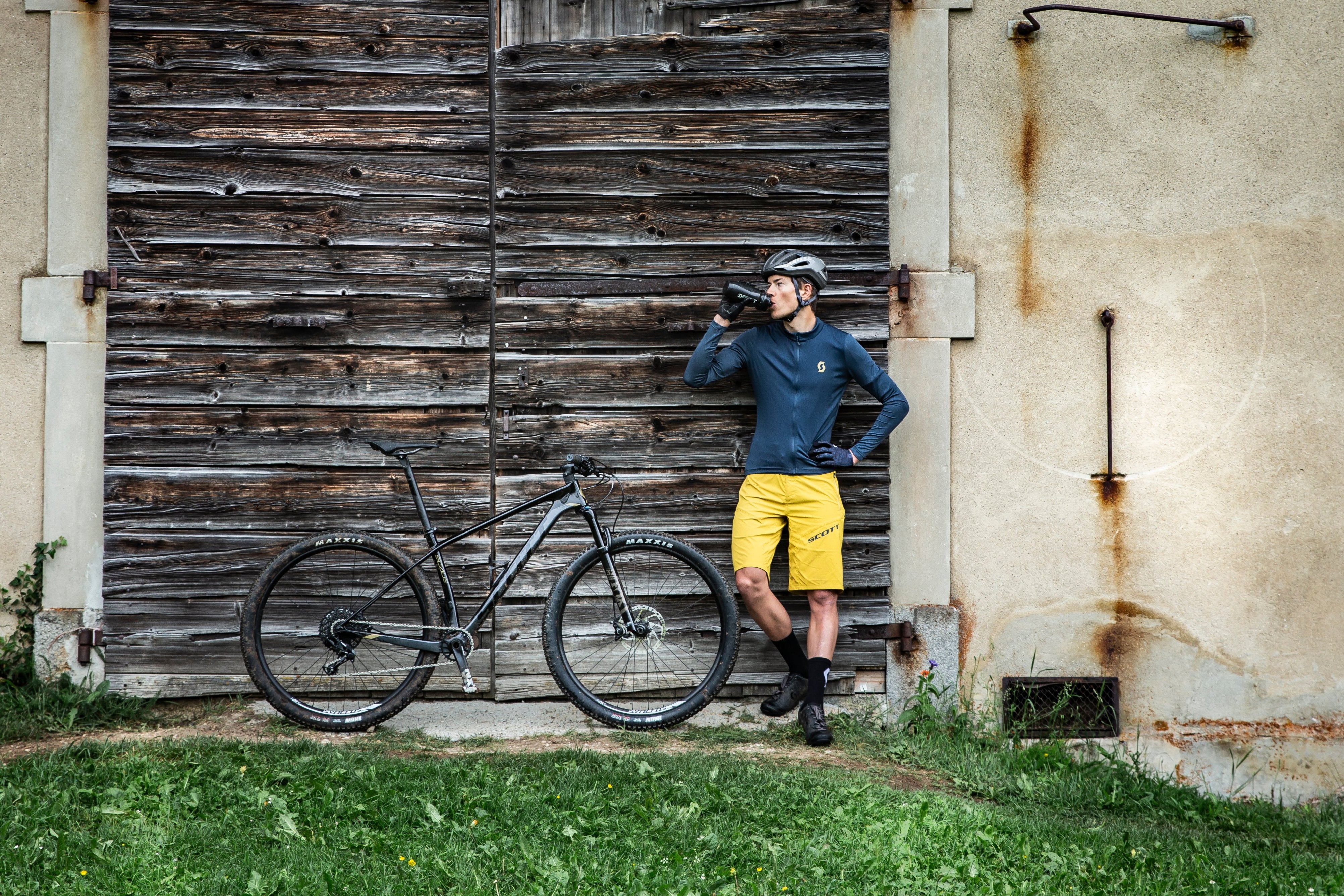 A man taking a break from riding his Scott Scale Mountain bike, while leaning against a building.