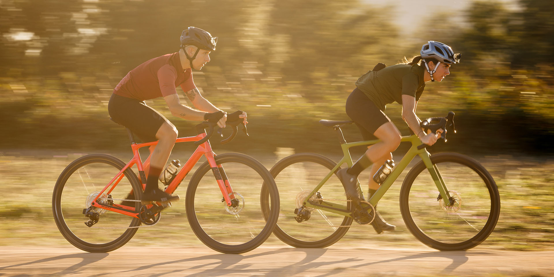 A man and a women riding the Scott Addict Gravel bike, one is painted orange, and one is green. they are riding the bikes on a dirt road by some trees. 