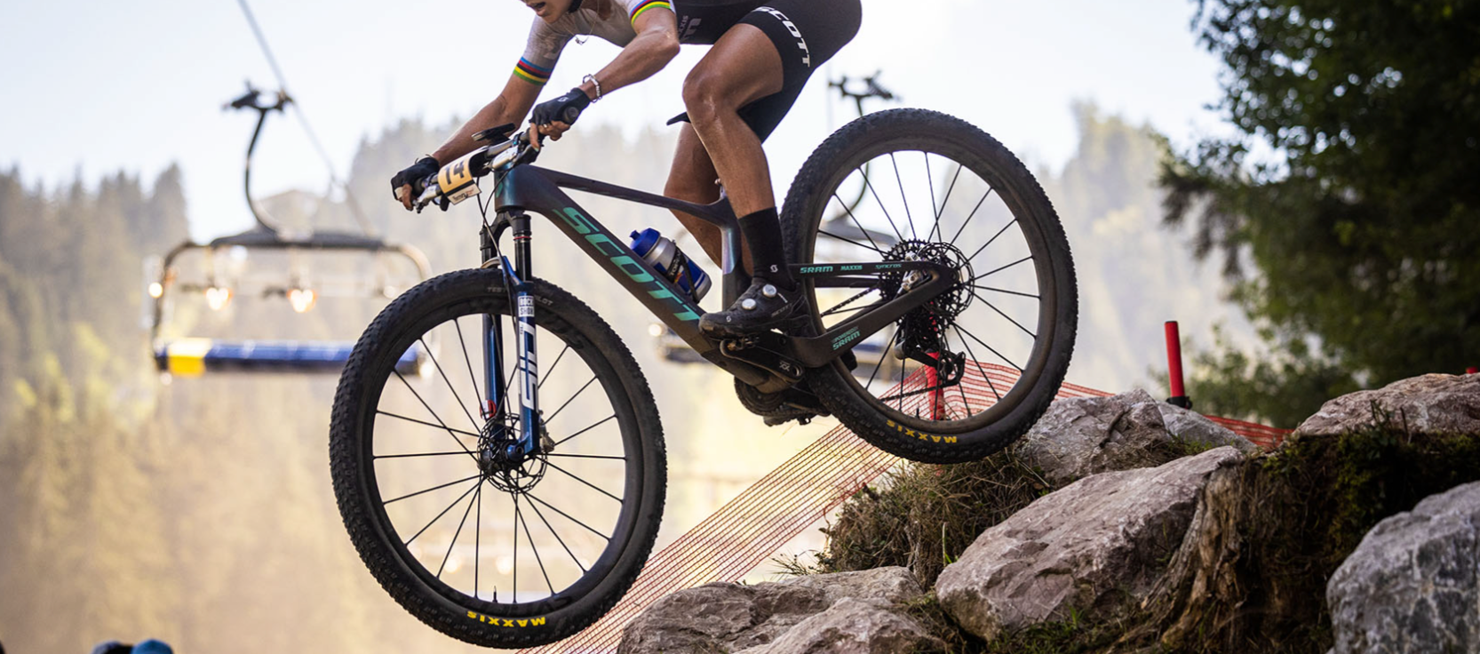 A person riding the Scott Spark RC mountain bike over some rocks in a forsest, with a chairlift in the background. 