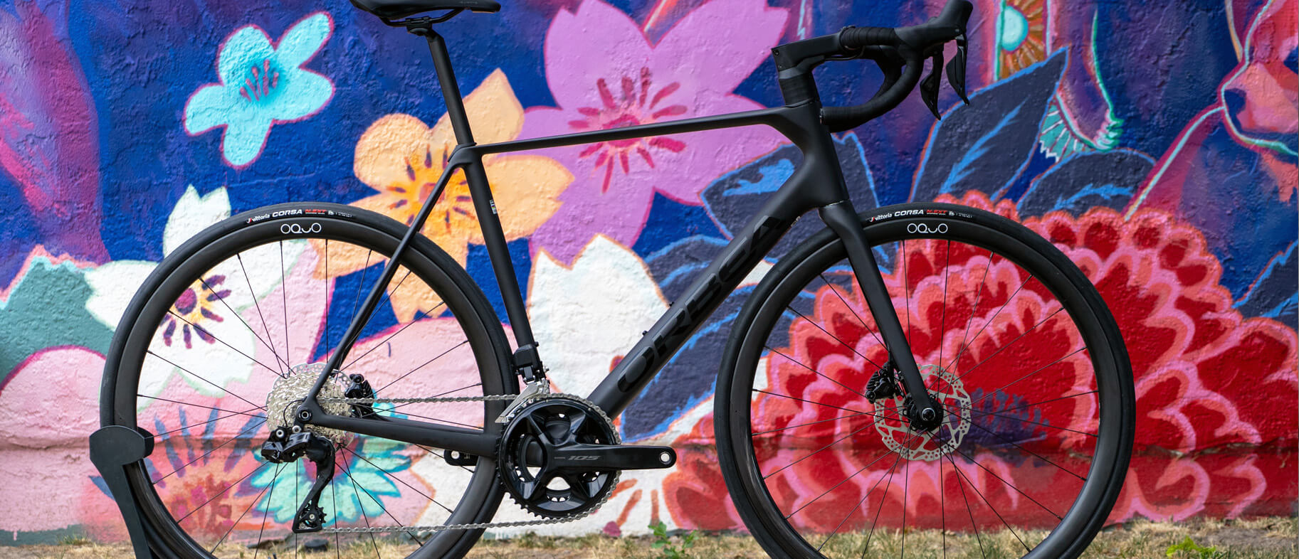 A black Orbea Orca Road bike against a colorfully painted wall.