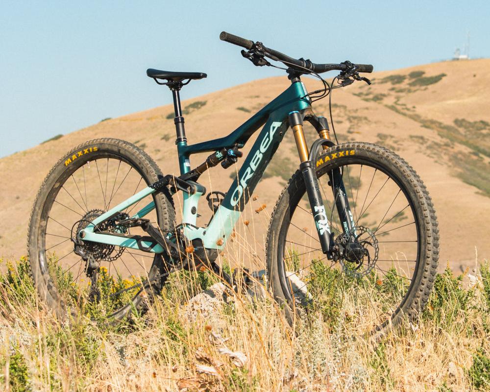 An Orbea Rise E-Mountain bike with teal and green paint, placed at the top of a hill with some mountains in the background.