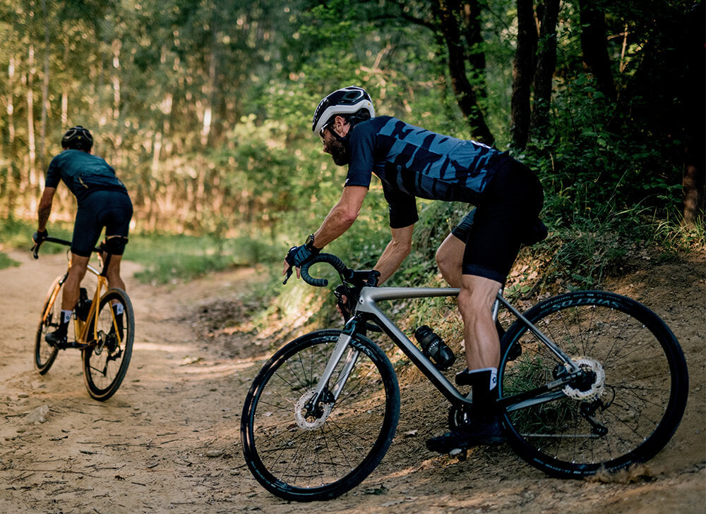 Two men riding the Scott addict gravel bike in a forest on a dirt road. One bike is gray, and one is yellow. 