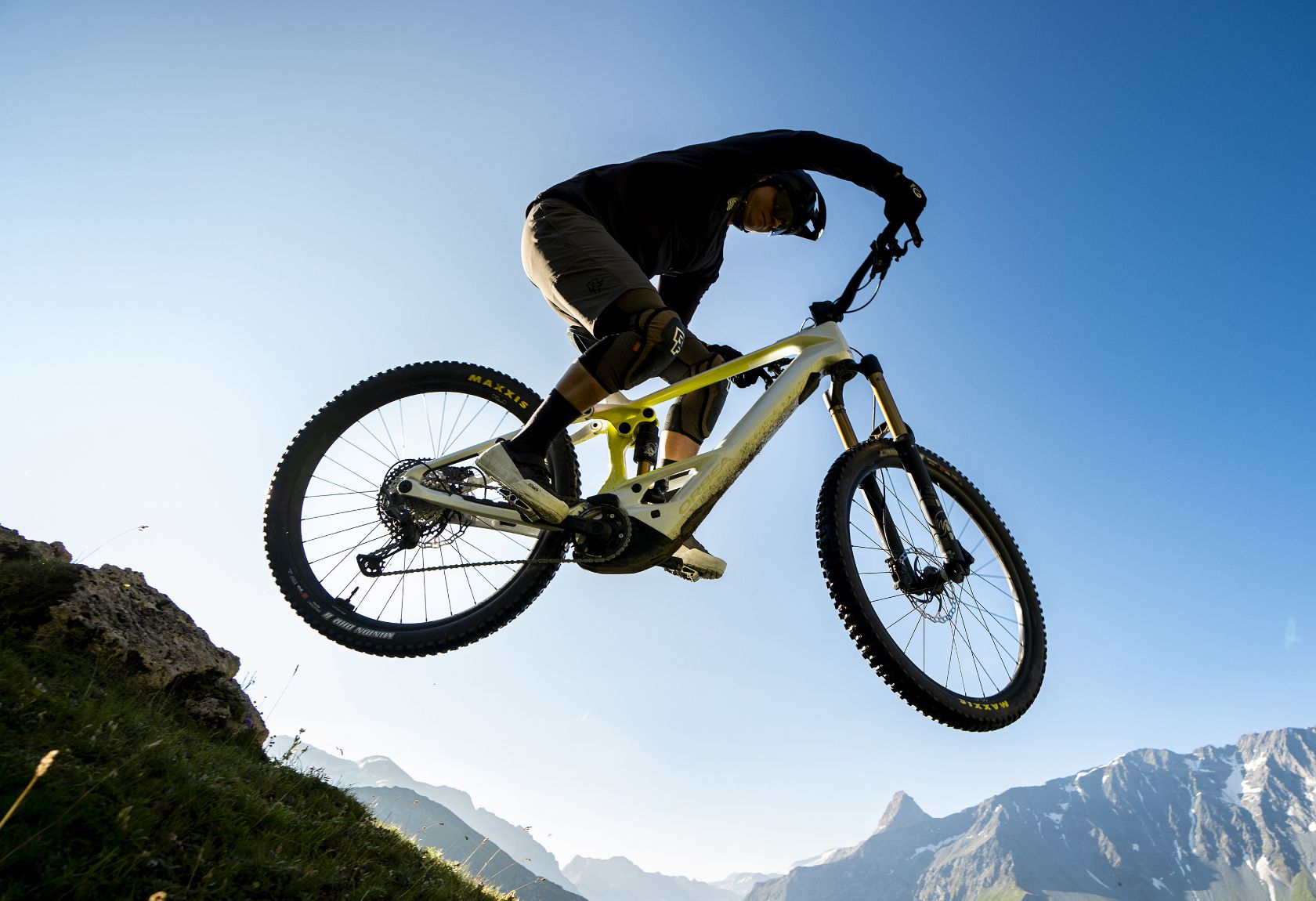 A man riding the Orbea Wild E-mountian bike with white paint, going off a jump with some mountains in the background. 